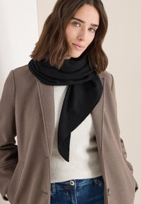 Bufanda negra con una sutil textura y brillo, drapeada alrededor del cuello, combinada con un suéter de canalé en un color claro y un abrigo marrón.