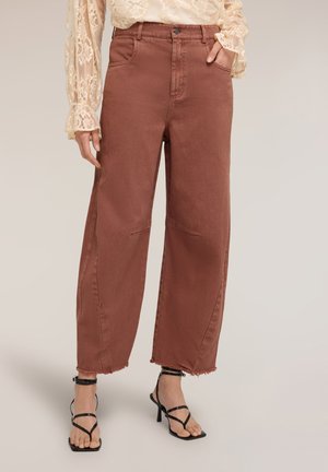 Kobieta w wysokich, szerokich spodniach w kolorze rdzawego brązu z surowymi zakończeniami, zestawionych z czarnymi sandałami na obcasie z paskami oraz kremową koronkową bluzką.