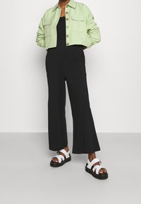 Femme portant une combinaison noire à jambes larges, une veste verte claire à boutons coupée, et des sandales blanches à plateforme avec de grosses lanières.