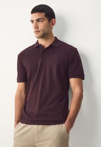 Bordo spalvos polo marškinėliai iš lengvos medžiagos, su klasikiniais apykakle, dviejų mygtukų užsegimu ir subtiliu logotipu ant krūtinės. Lygios tekstūros.