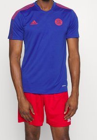 Modrý sportovní dres s červenými akcenty, s krátkými rukávy, výstřihem do V a logem FC Bayern Mnichov. V kombinaci s červenými šortkami.