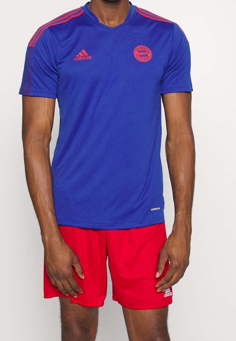 Modrý sportovní dres s červenými akcenty, s krátkými rukávy, výstřihem do V a logem FC Bayern Mnichov. V kombinaci s červenými šortkami.