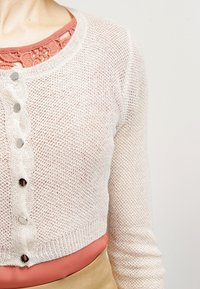 Lehký, otevřený krémový cardigan s dlouhými rukávy a dekorativními knoflíky. Nosený přes korálový top s krajkovým detailem na výstřihu.