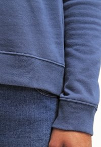 Felpa blu aderente con tessuto testurizzato, polsini e orlo a coste. Indossata sopra jeans in denim, che mostrano un look casual a strati.