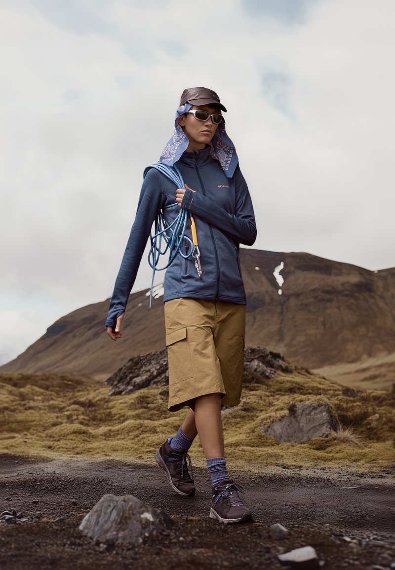 Persoon in een marineblauwe ritsjack, kaki cargoshorts en bruine wandelschoenen, terwijl hij klimtouwen vasthoudt met een gemusterde bandana en pet.