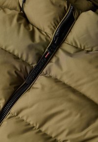 Nahaufnahme einer olivgrünen Steppjacke mit einem schwarzen, teilweise geöffneten Superdry-Reißverschluss, die die wattierte Stoffstruktur zeigt.