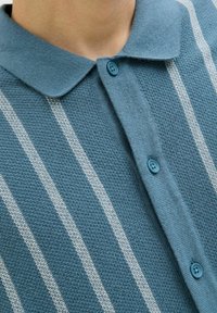 Pull turquoise avec un design col, comportant des rayures verticales blanches, un tissu texturé et une fermeture à bouton bleue au cou.