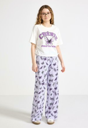 Ung kvinde med briller iført hvid T-shirt med lilla sommerfugl og teksten "CHÉRIE" samt lyse lilla vide bukser med sommerfuglemønster.