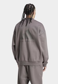 Grå sweatshirt med ribbad nederkant och ärmslut, med en stor matt svart Adidas-logotyp och texten "BASKETBALL" nedanför på baksidan.