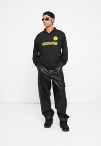 Schwarzes Langarmshirt mit gelbem "ANACHE"-Logo, kombiniert mit lockeren schwarzen Cargo-Hosen aus Kunstleder und schwarzen Sneakers. Strukturierte Oberflächen sichtbar.