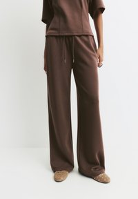 Persoană îmbrăcată cu pantaloni maronii largi, cu șnur, și pantofi slip-on maronii cu detalii din metal, stând în fața unui fundal alb.