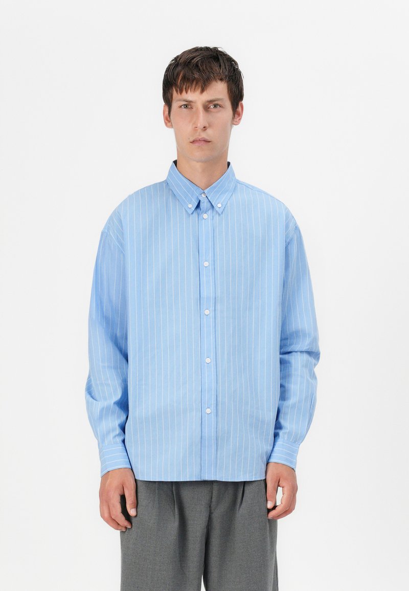 Chemise boutonnée bleu clair avec des rayures verticales blanches, dotée d'un col pointu et de manches longues, assortie à un pantalon gris.