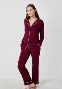 Bordeaux pyjama set met een langlopende knoopsluiting bovenstuk met witte bies en bijpassende broek met zijzakken. Zacht, glad materiaal.