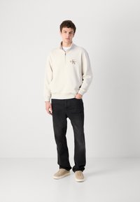 Felpa leggera beige con zip e logo, abbinata a jeans scuri a gamba dritta e sneakers beige, su uno sfondo grigio uniforme.