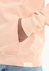 Jasnoróżowy sweatshirt o miękkiej teksturze, z prążkowanymi mankietami, boczną kieszenią i metką przy hemie z napisem "Scotch & Soda."