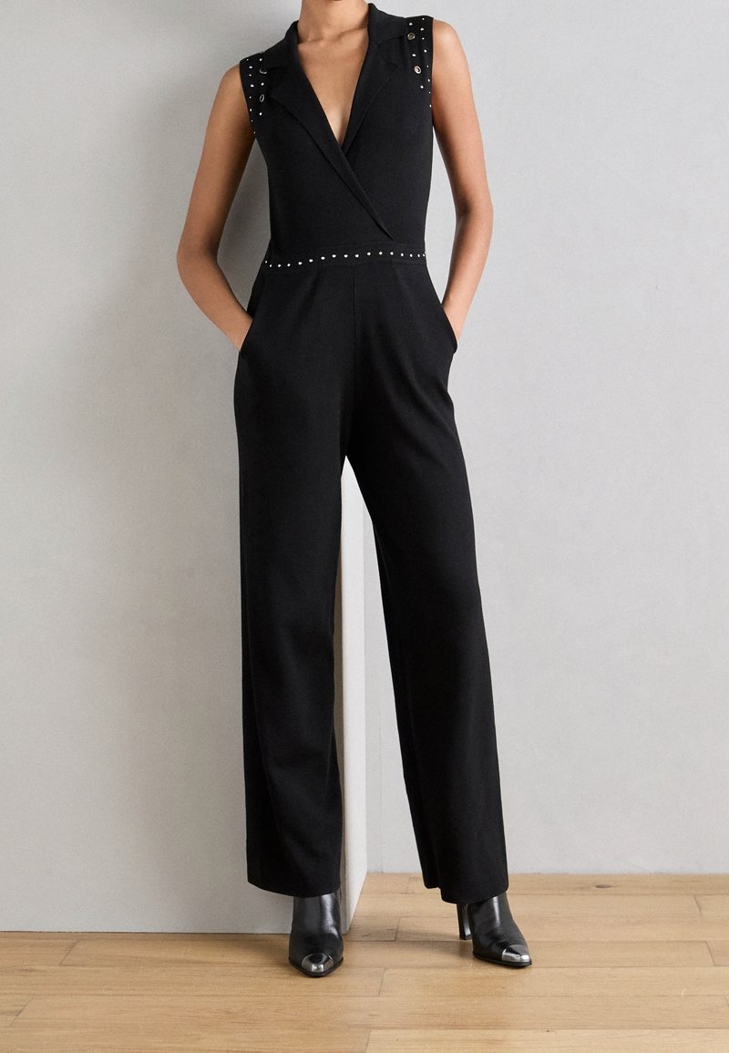 Femme portant une combinaison noire sans manches avec des détails cloutés et des jambes larges, associée à des bottines noires à bout pointu, se tenant contre un mur gris.