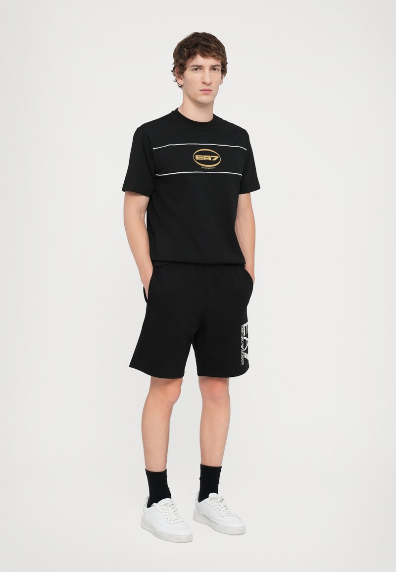 Juodas marškinėlis su auksiniu logotipu ir baltais dryžiais, derinamas su juodomis šortais, turinčiais baltą grafiką. Baltos sportbačiai ir juodos kojinės.