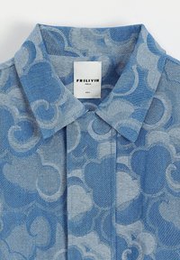 Chemise en denim bleu avec un motif tourbillonnant bleu clair, col pointu et une étiquette indiquant "FRILIVIN, PARIS.FR, TAILLE S." Texture lisse.