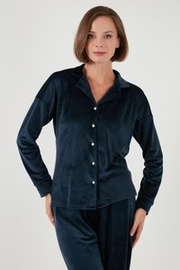 Vrouw draagt een marineblauw fluwelen pyjamapak met lange mouwen en witte knopen, staand tegen een effen lichte achtergrond.