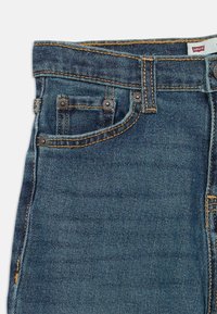Jeans de ganga azul com lavagem clara, apresentando costura em contraste, um design clássico de cinco bolsos e rebites de metal nos bolsos frontais.