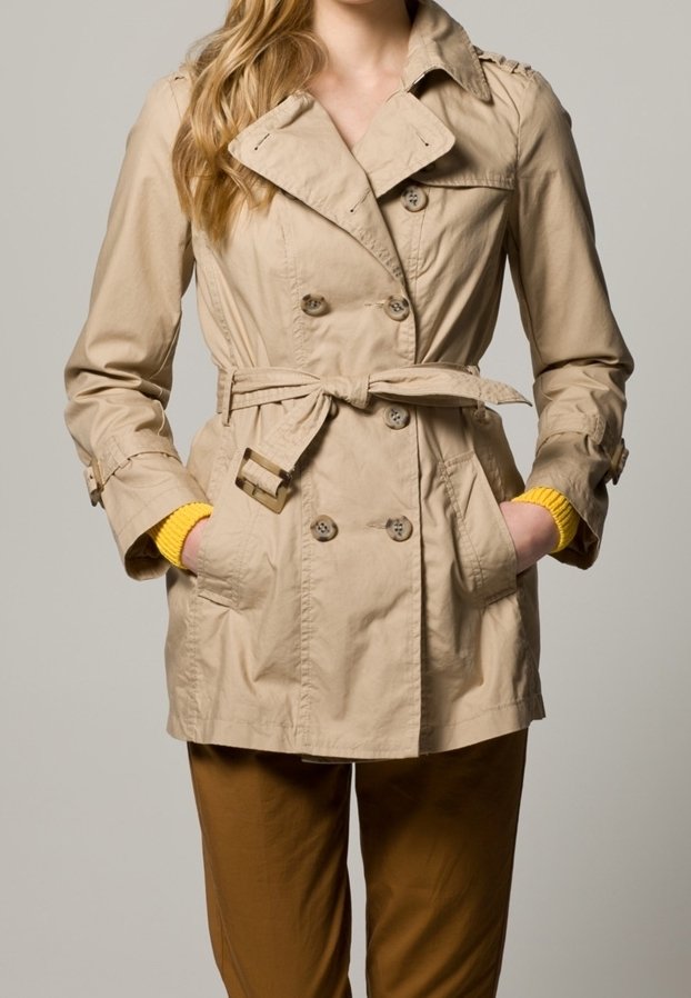 Beige trenchcoat med dubbelknäppta knappar, bälte i midjan och axelklaffar. Har sidofickor och en slät yta.