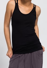 Femme portant un débardeur noir sans manches à double bretelle et un pantalon gris ample, debout avec une main dans la poche.