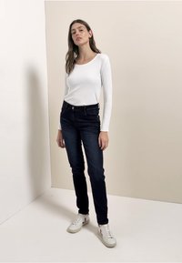 Valkoinen pitkähihainen paita, yhdistetty tummansinisiin kapeisiin farkkuihin ja valkoisiin tennareihin. Taustalla vaalea neutraali sävy.