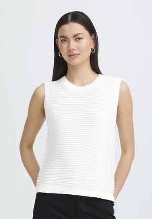 Sirge tumedate juustega naine, kes kannab hõbedasi rõngaskõrvarõngaid ja valget varrukateta soonikutega kootud pusa, taustal hele ühtlane taust.