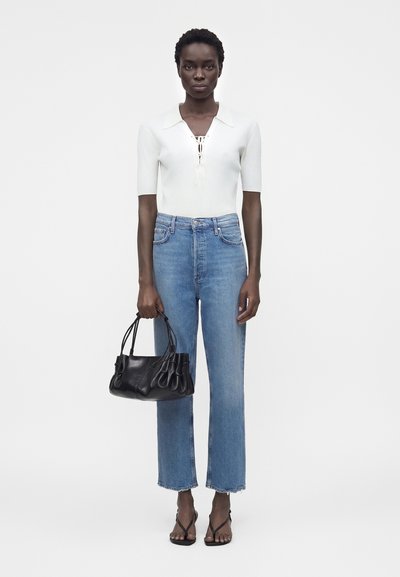 Γυναίκα που φοράει λευκό κοντομάνικο μπλουζάκι, μπλε straight-leg τζιν, μαύρα σανδάλια, κρατάει μαύρη δερμάτινη τσάντα, στέκεται μπροστά σε απλό φόντο.