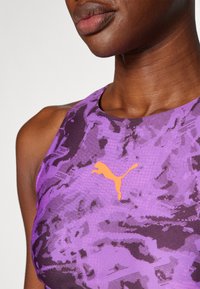 Violetti urheilu-toppi, jossa on teksturoitu muotoilu. Rinnassa on tyylitelty oranssi Puma-logo ja korkea kaulus.