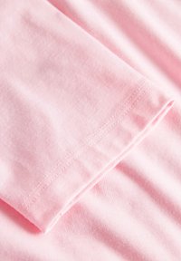 Kortärmad, ljusrosa bomullströja i närbild. Slät textur, synlig sömnad på den rullade ärmen, vikt tygkant.