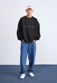 Felpa nera con il testo "Tutto lavoro e niente svago" in teal, abbinata a jeans blu larghi e sneakers bianche. Il modello indossa un cappellino beige.