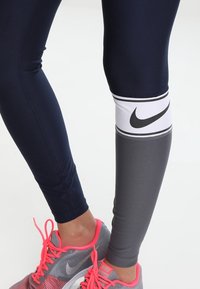 Marinblå träningsleggings med en vit Nike-logotyp och grå panel på underbenet. Matchas med grå sneakers med rosa inslag.