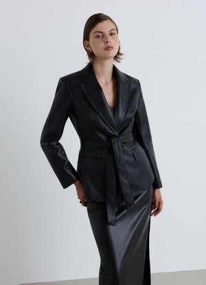 Donna che indossa un blazer in pelle nero con cintura e una lunga gonna abbinata, in piedi davanti a uno sfondo grigio chiaro uniforme.