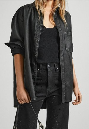 Schwarzes Denimhemd mit lockerem Schnitt, dezentem Glanz und durchgehender Knopfleiste, kombiniert mit schwarzen Jeans mit Nietenakzenten.