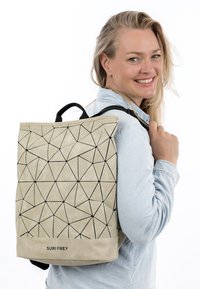 Nainen hymyilee katsellen olkapään yli, pukeutuneena vaaleaan farkkupaitaan ja beigeen, mustilla remmeillä varustettuun geometrisesti kuvioituun reppuun.