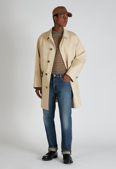 Manteau beige avec de grands boutons, pull en laine marron, jean bleu et chaussures en cuir noires. Le mannequin porte également une casquette marron.