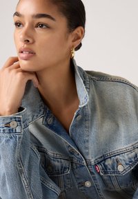 Νεαρή γυναίκα με χρυσούς κρίκους στα αυτιά, που φοράει μπουφάν από ξεθωριασμένο μπλε τζιν, στηρίζοντας το πηγούνι της στο χέρι της, κοιτάζοντας προς τα πλάγια.