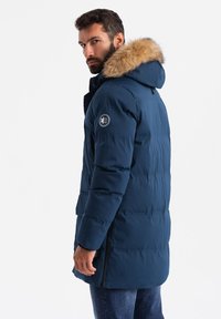 Mörkblå puffjacka med en huva fodrad med päls, quiltat mönster, två siddragkedjor och en logotypapplikation på vänster ärm.