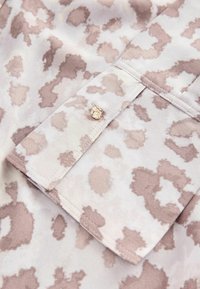 Leopardenmusterhemd aus weichem Material, mit beigen und blassen Flecken. Details umfassen einen Knopfakzent und einen umgeschlagenen Kragen. Glatte Textur.