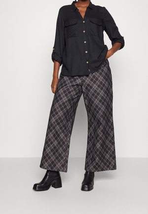 Person wearing black button-up shirt, gray and black wide-leg plaid pants, and black heeled ankle boots, standing with one hand in pocket.