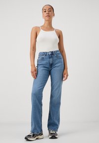 Top de alças branco canelado e jeans de cintura alta azul claro, com um corte descontraído e perna reta. O modelo usa ténis pretos e brancos.