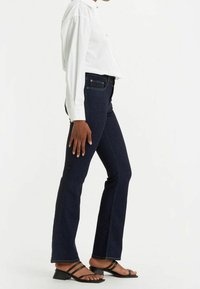 Pantalones vaqueros de campana en azul oscuro con una cintura alta, corte recto y sutiles detalles de costura, combinados con una camisa blanca de botones y sandalias negras.