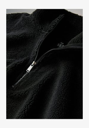 Mustast fliisist pullover, millel on kõrge krae ja veerandlukk, pehme tekstuuriga pind ja metallist lukk. Silt on osaliselt nähtav.