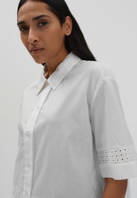 White button-up shirt with a collar and short sleeves, featuring embroidered cut-out detailing at the sleeves and a smooth, textured fabric.