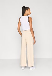 Fehér ujjatlan felső fekete szegéllyel, bézs, bőszárú nadrággal párosítva. A modell hátulról látható, fehér edzőcipőt visel.