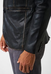 Schwarze Lederblazer mit tailliertem Design, sichtbaren Nähten, einem einknopfigen Verschluss und drei Knopf-Akzenten an den Ärmeln.
