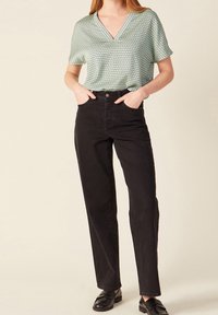 Donna che indossa jeans dritti neri a vita alta, top a maniche corte con scollatura a V di un verde chiaro e pattern, e mocassini neri, con le mani in tasca.