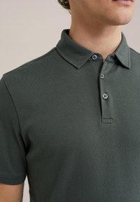Polo de color verde oscuro hecho de un tejido texturizado, con un cuello tipo camisa, tapeta de tres botones y detalles de costura visibles.