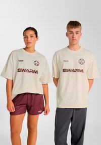 Beige oversized t-shirts med logo, kombineret med bordeaux shorts til venstre og grå træningsbukser til højre, hvilket fremhæver et sportigt design.